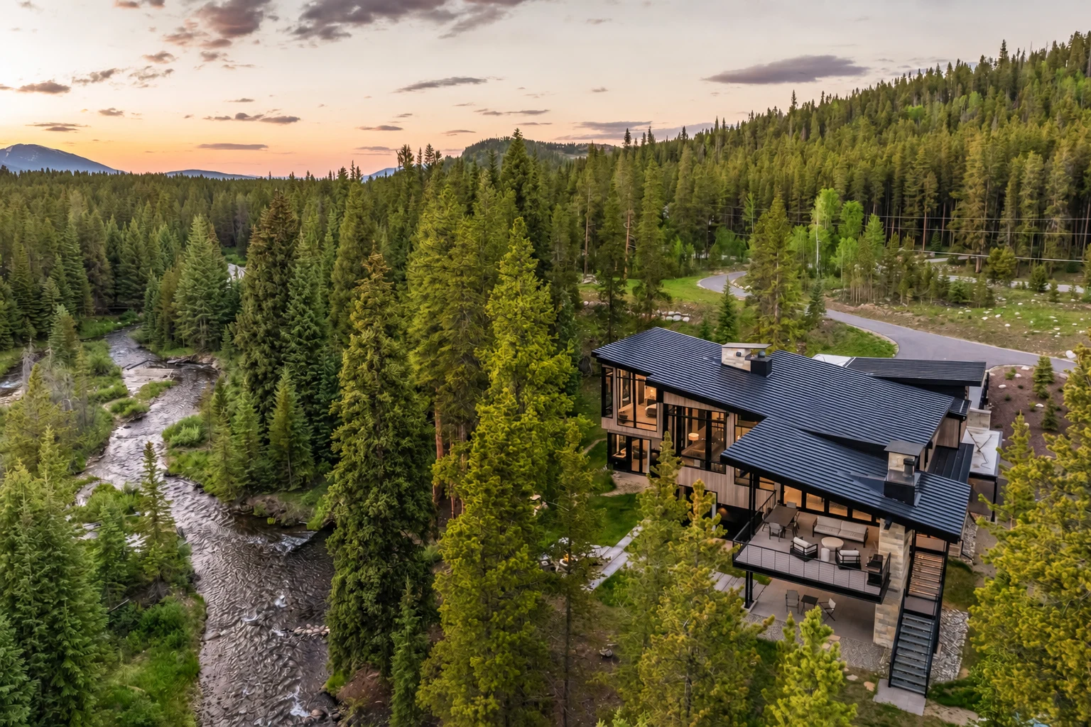 Aerial view of a modern mountain home beside a river and pine forest at sunset